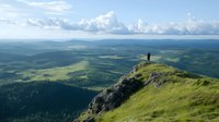 Lone hiker silhouette standing on mountain peak overlooking vast green valley landscape with rolling hills and dramatic clouds in summer wilderness.