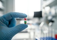 Hand in blue glove holding small test tube with colorful beads in laboratory setting, used for medical research or diagnostic testing.