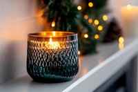 Glowing candle in textured blue glass holder on white windowsill with blurred Christmas tree and warm bokeh lights creating cozy holiday atmosphere.