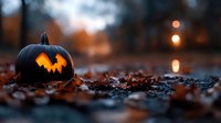 Glowing carved pumpkin with jagged smile on autumn ground covered with fallen leaves at dusk, creating spooky Halloween atmosphere with bokeh lights in background.
