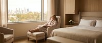 Young woman in bathrobe relaxing in luxury hotel room with city view. Modern hospitality interior design for travel and wellness lifestyle concepts.