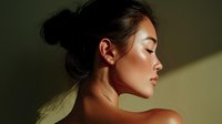 Young Asian woman with natural makeup in profile view against dark background, eyes closed, showing bare shoulders and glowing skin in dramatic lighting.