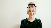 Young woman with blonde hair and fair complexion wearing dark green velvet top and burgundy bow headpiece, smiling at camera against neutral background for portrait and lifestyle use.