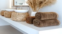 Folded brown towels and woven storage baskets on white bathroom shelf with dried wheat decor, creating minimal spa-like atmosphere for modern interior design.