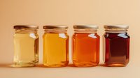 Different varieties of honey in glass jars showing color gradient from light to dark amber, arranged in row against beige background.