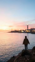 Person standing on rocky shore gazing at sunset over waterfront city skyline with illuminated lighthouse and blurred urban lights reflecting on calm water.