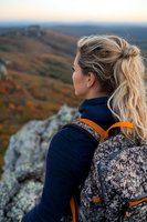 Young blonde woman in navy sweater with backpack enjoying scenic mountain view during autumn sunset, contemplating nature from rocky overlook.