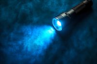 Black tactical flashlight beam cutting through blue smoke or fog creating atmospheric light rays and particles in dark background.