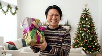 Happy Asian man holding wrapped Christmas gift with purple ribbon in festive living room decorated with tree and garland during holiday celebration.