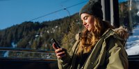 Young woman in winter clothing checking smartphone on ski lift with snowy mountain landscape in background. Perfect for travel and winter sports content.