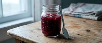 Homemade berry jam in glass jar with spoon on rustic wooden table near window. Fresh fruit preserve for breakfast, baking and cooking recipes.