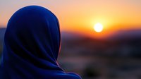 Woman wearing dark blue hijab silhouetted against vibrant orange sunset sky with glowing sun on horizon, peaceful contemplative moment captured in soft focus.