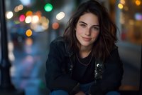 Young woman with long dark hair and fair complexion wearing black jacket poses outdoors at night with colorful bokeh city lights in blurred background, suitable for lifestyle and portrait use.
