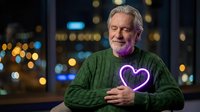 Senior Caucasian man with gray beard holding glowing purple heart light against city night bokeh background. Concept of love, romance, and emotional connection.