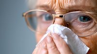 Senior woman with glasses blowing nose into tissue during cold or flu season, close-up view showing facial wrinkles and eyewear against blurred background.