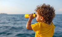 Young mixed-race child with curly hair wearing yellow t-shirt looks through binoculars at sea horizon during sunset, exploring nature and adventure concept.