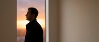 Young professional man silhouette standing by window during golden hour sunset, contemplating future opportunities in modern office space.