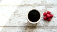 Overhead view of steaming coffee cup with fresh raspberries on rustic whitewashed wooden surface, ideal for breakfast and beverage content.