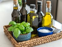 Assorted olive oil bottles and balsamic vinegar arranged on woven tray with fresh basil plant and sea salt in ceramic bowl, Mediterranean cooking ingredients.