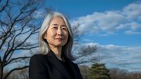 Confident mature Asian woman with long silver hair wearing black business attire stands outdoors against dramatic blue sky with white clouds and bare winter trees.