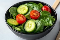 Fresh spinach salad with sliced cucumbers and cherry tomatoes in black bowl. Healthy green leafy vegetables for nutritious meal preparation and diet.