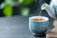 Steaming hot tea being poured from traditional ceramic teapot into decorative Asian cup with blue pattern design on blurred natural background. Calming drink moment.