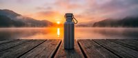 Thermos bottle on wooden dock at sunrise over misty lake with mountains, ideal for beverage, outdoor adventure, and travel lifestyle marketing.