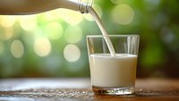 Fresh milk pouring into glass on wooden surface with natural green bokeh background for healthy breakfast and dairy product concepts.
