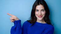 Young Caucasian woman with dark hair wearing bright blue sweater pointing finger sideways while smiling cheerfully against blue background. Suitable for advertising and lifestyle content.