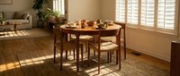 Warm wooden dining set with round table and chairs in bright sunlit room with shuttered windows, hardwood floors, and natural home decor for interior design projects.