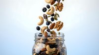 Mixed nuts and dried fruits falling into glass jar on white background for healthy snacking and nutrition concepts.