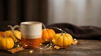 Warm mug of amber cider surrounded by small decorative gourds, dried leaves, and string lights on wooden surface for autumn beverage and seasonal home decor projects.