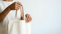 Woman with medium skin tone holding empty natural canvas tote bag, wearing white casual shirt. Suitable for eco-friendly shopping, sustainable lifestyle, and retail branding campaigns.