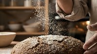 Baker dusting flour onto fresh dough in rustic kitchen workshop. Artisan bread making process with flour particles suspended in air.