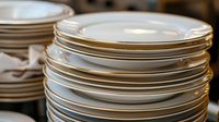 Stack of white porcelain plates with gold trim arranged in piles on dark background, elegant dinnerware for restaurant and catering service setup.