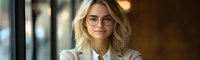 Young professional woman with blonde wavy hair and glasses smiling confidently in modern office setting, wearing beige blazer and white blouse, natural window lighting.