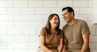 Young couple, Caucasian and Hispanic ethnicity, smiling and laughing together against white subway tile wall. Casual lifestyle moment capturing joy and connection between partners in relaxed home setting.