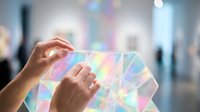 Hands holding transparent holographic geometric shapes with rainbow prism effects in modern gallery space for futuristic technology and augmented reality concepts.