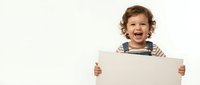 Happy toddler boy with curly brown hair holding blank white sign on beige background. Cheerful child in striped shirt and overalls displaying empty poster.