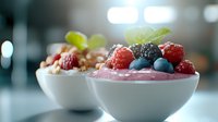 Fresh berry smoothie bowl topped with raspberries, blackberries, blueberries, granola, and mint leaves on blurred kitchen background.