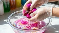 Hands in gloves washing vibrant pink sponge in glass bowl with soapy water for cleaning and household maintenance tasks.