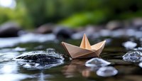 Paper boat floating on water surface among stones, with soft bokeh background and natural lighting creating dreamy atmosphere. Childhood memories concept.