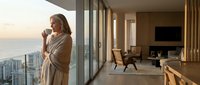 Woman in bathrobe drinking coffee on luxury apartment balcony with ocean view at sunrise, modern interior design with floor-to-ceiling windows and fireplace.