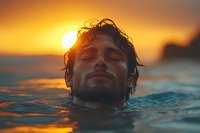 Young man swimming in ocean water at sunset with eyes closed, peaceful expression, wet hair backlit by golden sun, waves reflecting orange sky.