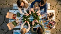 Family gathering around table making Christmas wreaths together with festive decorations and craft supplies on outdoor stone patio during holiday season.