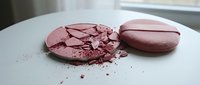 Broken pink makeup powder compact with crumbs scattered on white surface, cosmetic accident damage for beauty and skincare concepts.