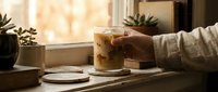 Person with light skin tone holding iced coffee drink at wooden desk with notebook, plant, and warm sunlight streaming through window for beverage and lifestyle content.