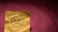 Yellow sticky note with hand-drawn sad face emoji on burgundy surface expressing disappointment, negativity, or unhappy emotions for office communication.
