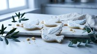 White dove-shaped cookies with decorative icing on marble surface with eucalyptus branches in bright kitchen setting for baking and celebration themes.