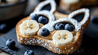 Playful breakfast toast shaped like bunny face with banana slices for eyes, fresh blueberries and powdered sugar on dark background. Creative food for kids.